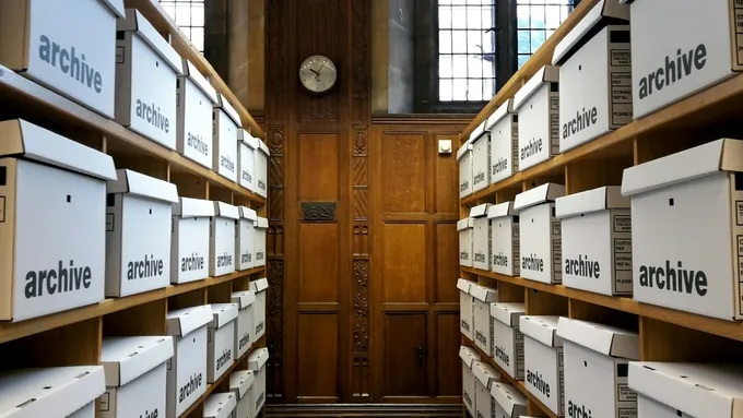 Valkoisia archive-tekstillä merkittyjä arkistolaatikoita puuhyllyillä kapean käytävän molemmin puolin, taustalla puupaneloitu seinä ja kello.