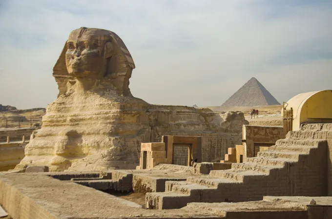 Suuri Sfinksi edestä kuvattuna kivisten raunioiden takana, oikealla taustalla pyramidi utuisen taivaan alla.