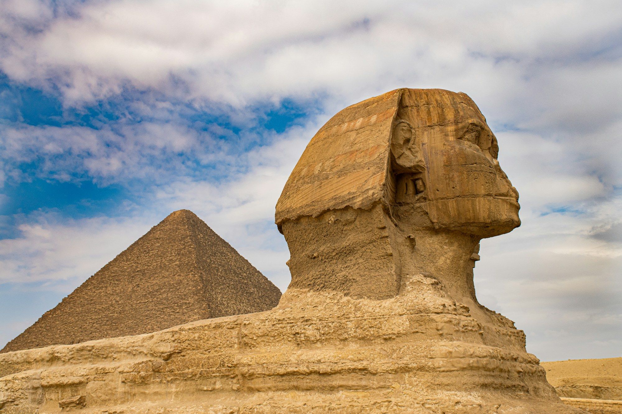 Gizan Suuri Sfinksi sivulta kuvattuna, kasvoista puuttuu nenä. Taustalla pyramidi pilvisen sinisen taivaan alla.