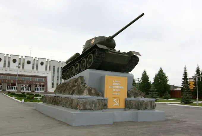 Jalustalle nostettu T-34-panssarivaunu, jonka takana on vaalea tehtaan päärakennus. Rakennuksen julkisivussa näkyy kyrillisiä kirjaimia.