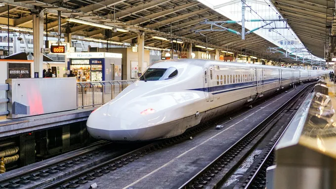 Valkoinen, sinijuovainen shinkansen-juna pitkine nokkineen seisoo katetun asemalaiturin vieressä.