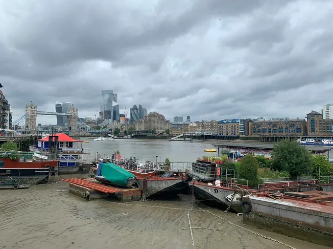 Asuntoveneitä ja proomuja laskuveden paljastamalla liejulla Thamesin rannassa, taustalla Tower Bridge ja Cityn pilvenpiirtäjiä pilvisen taivaan alla