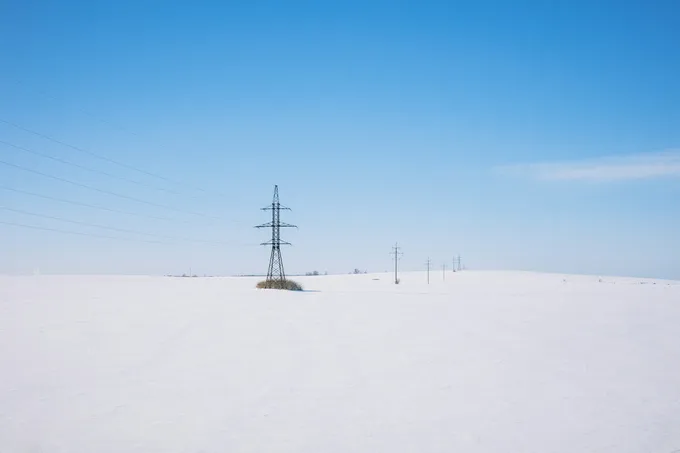 Sähkölinja ja ristikkopylväitä kulkee lumisen pellon poikki sinistä taivasta vasten.