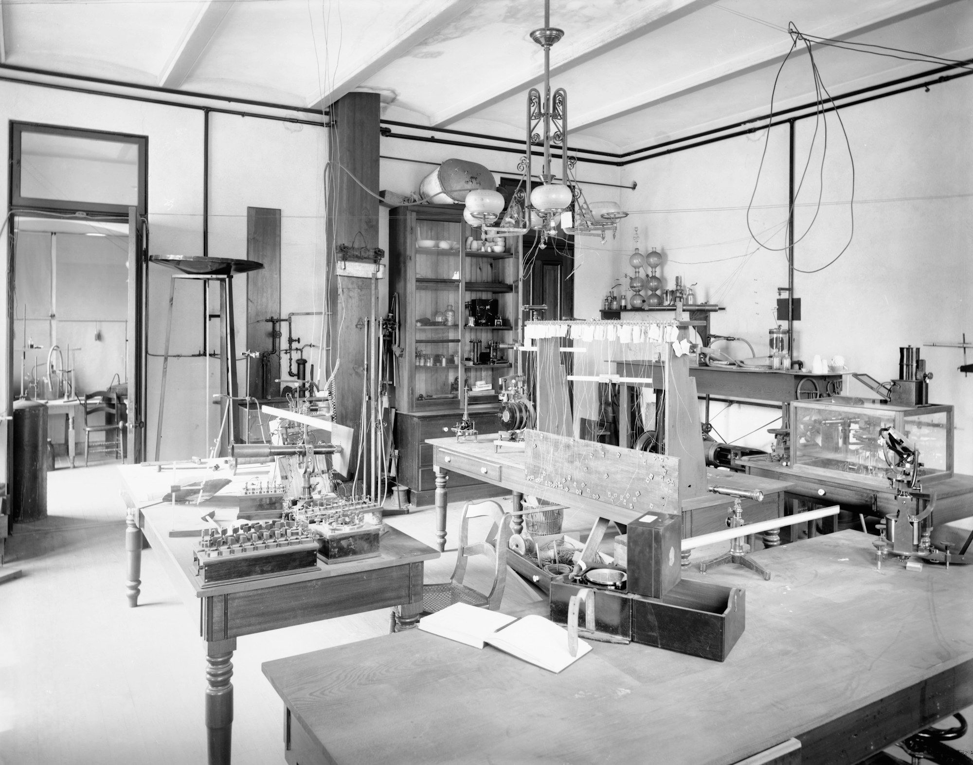 Mustavalkoinen kuva vanhasta laboratoriosta, jossa puupöydillä on sähkölaitteita, johtoja ja mittalaitteita, taustalla lasikaappi ja lasiastioita hyllyllä.