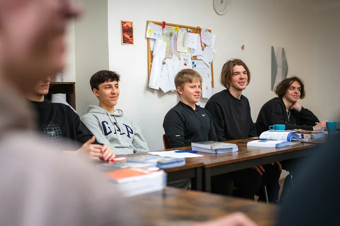 Nuoria miehiä istuu pöytien ääressä oppitunnilla, edessään oppikirjoja ja kahvikuppeja.
