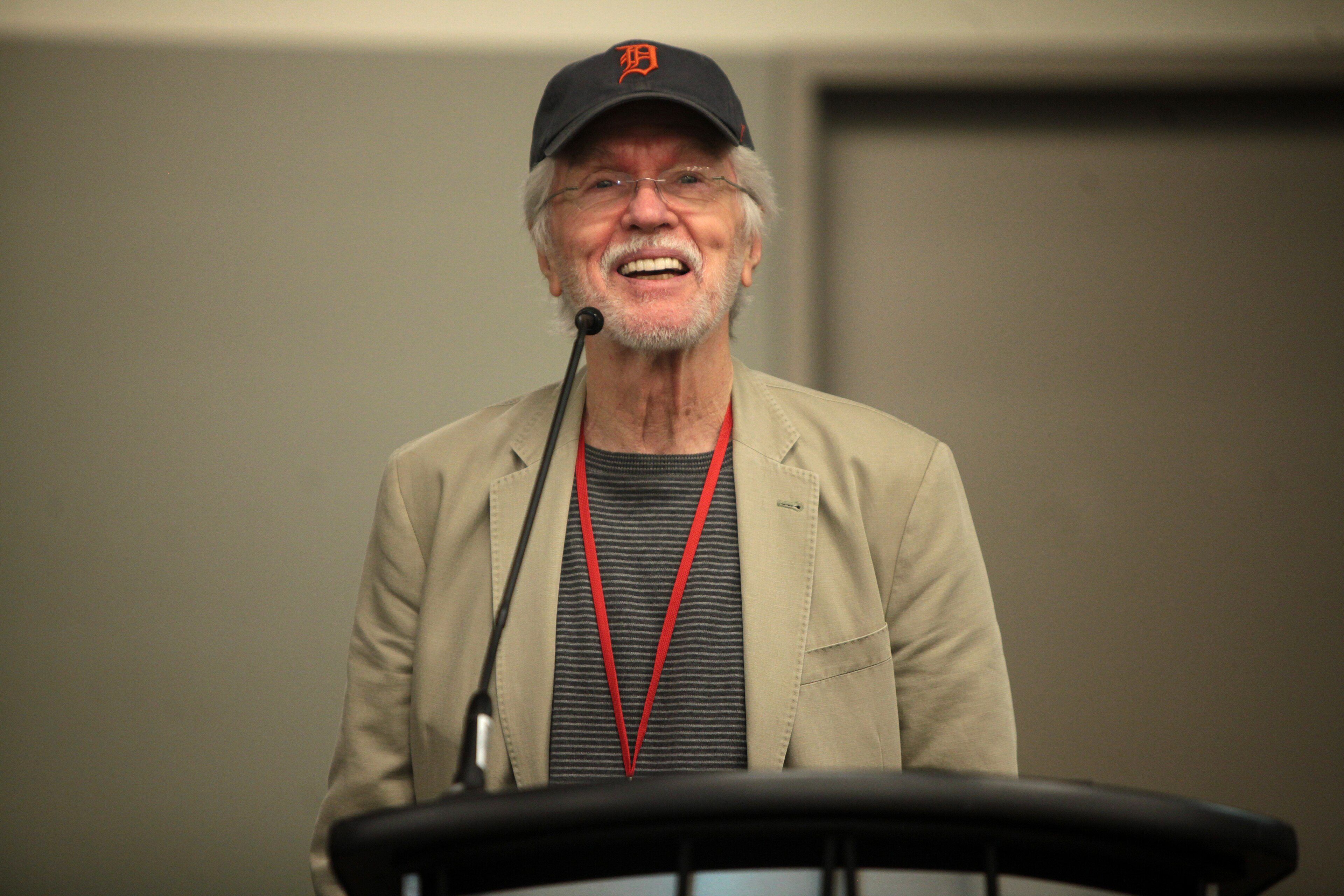 Harmaapartainen Tom Skerritt hymyilee lippalakki päässä ja beige pikkutakki yllä puhujapöntön mikrofonin edessä.
