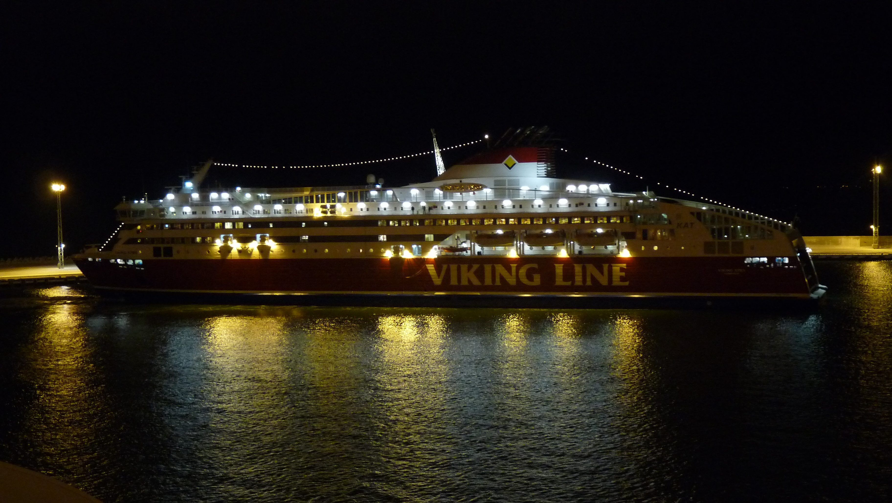 Valaistu punavalkoinen matkustajalaiva laiturissa yöllä. Laivan kyljessä lukee Viking Line, ja valot heijastuvat tummaan veteen.