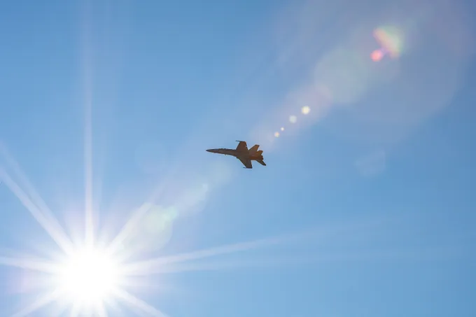Hornet-hävittäjän siluetti kirkkaalla sinisellä taivaalla, vasemmassa alakulmassa aurinko.